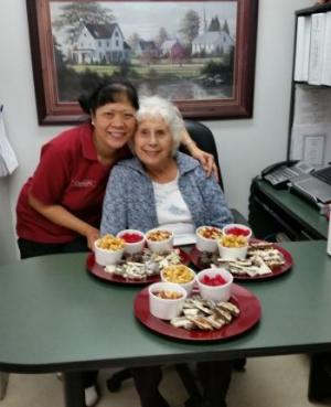 Erlinda leaning down with an arm over a female resident who is sitting at a table with three trays of food in front of her